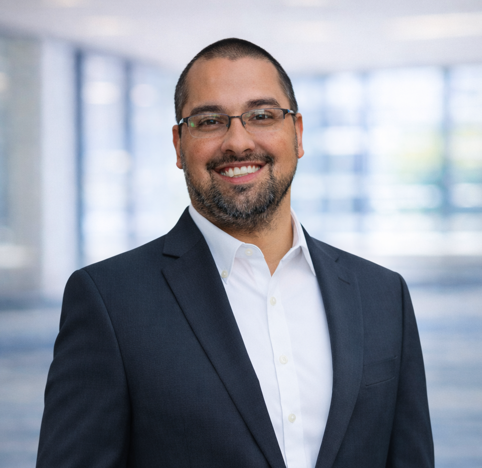 Smiling man in a checked shirt and dark blazer standing in front of green palm leaves